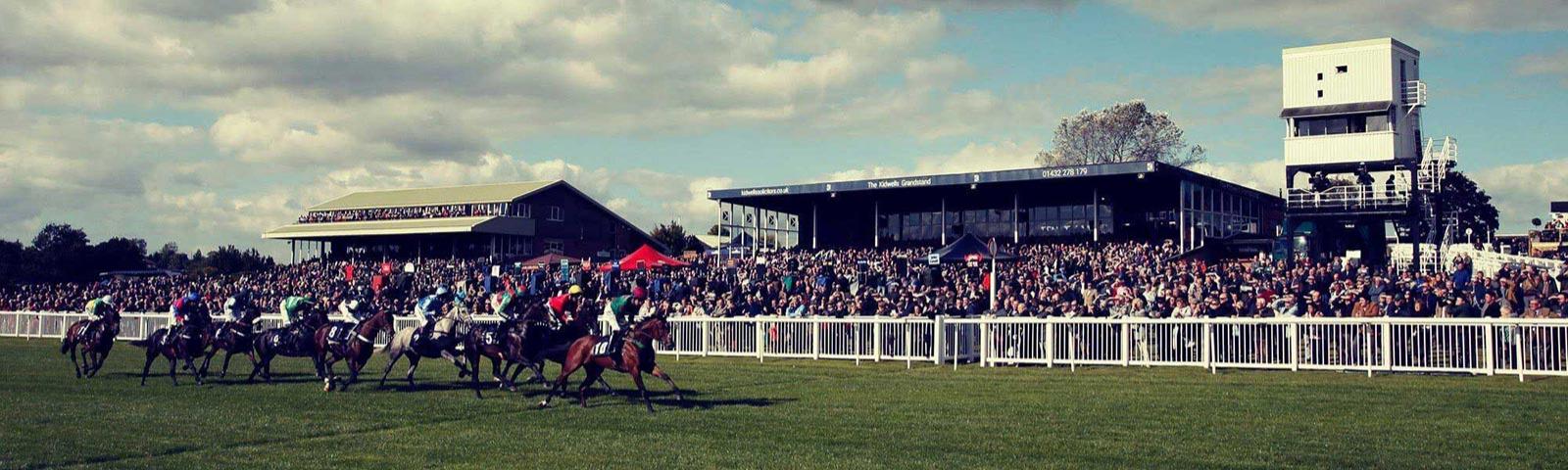 A raceday at Hereford Racecourse with crowds watching the jockeys race past.