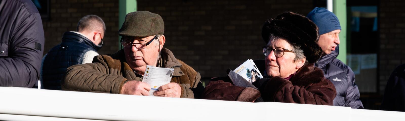 Two visitors to Hereford Races watching the parade ring while holding racecards